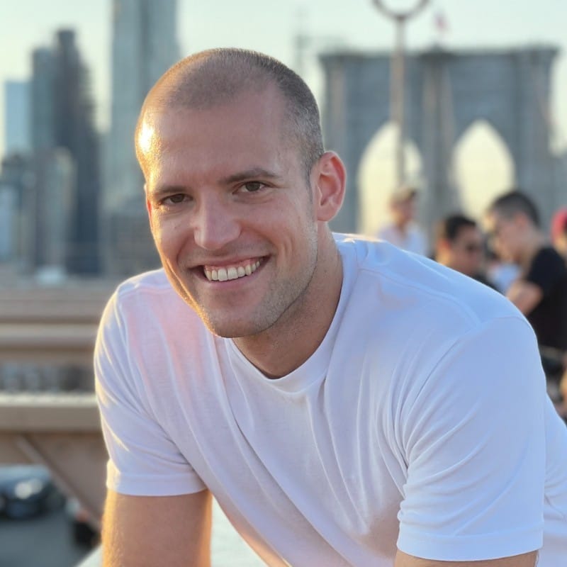 A man smiling while sitting on a bench on the brooklyn bridge.