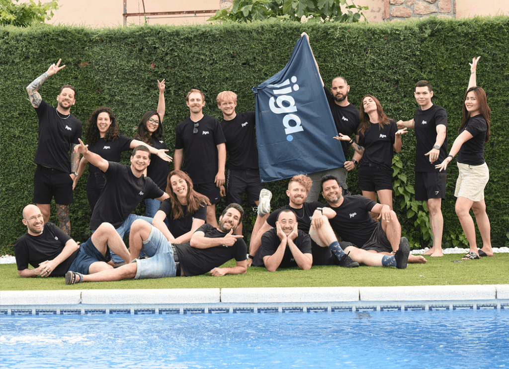 A group of people in matching shirts pose and smile for a photo by a pool, holding a flag with the word "jiga" in front of a hedge.