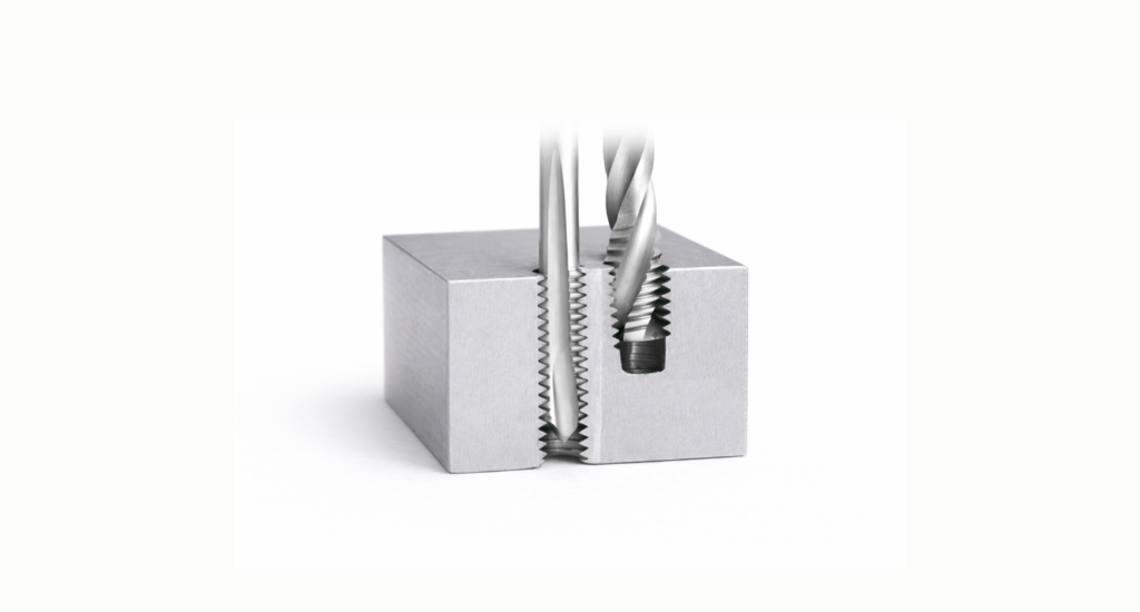 Metal block with a threaded hole is shown holding a screw and a corresponding tap tool side by side, illustrating the difference between tapped holes vs threaded holes during the threading process.