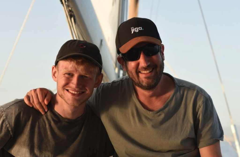 Two men wearing caps and casual shirts smile at the camera on a sailboat, with one man’s arm around the other’s shoulder.