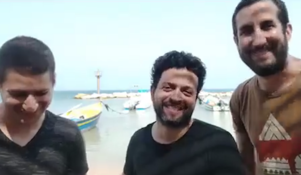 Three men standing and smiling in front of docked boats on a calm body of water under a clear sky.