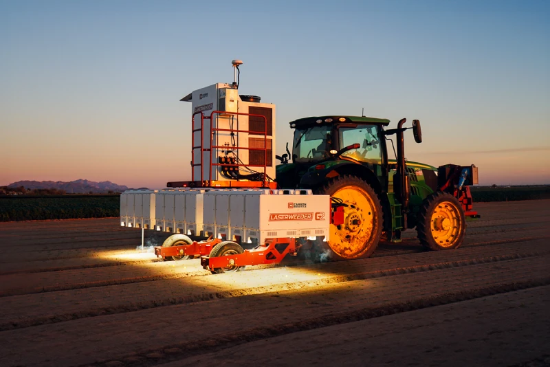 A tractor equipped with agricultural machinery and bright lights operates in a field at dusk or dawn, demonstrating the precision of custom parts sourcing for robotics in modern farming.