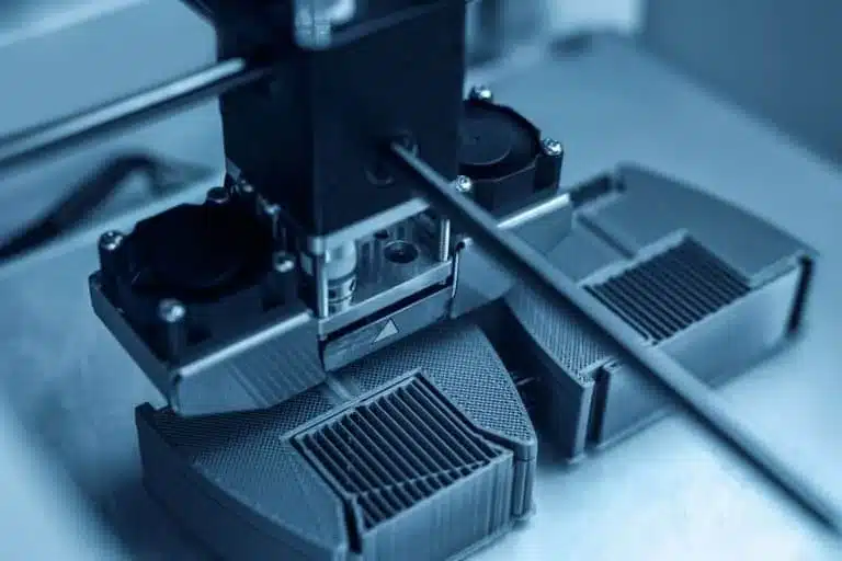 A 3D printer in operation, printing two black plastic objects with visible layered patterns on the print bed.