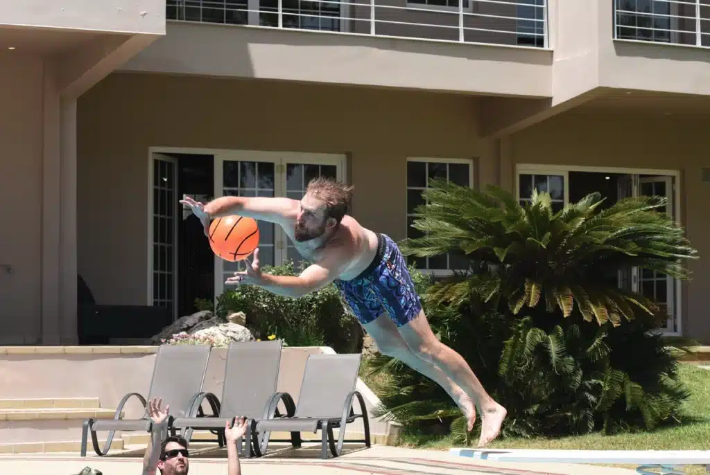 jiga executive in swim trunks leaps horizontally by a pool, reaching for an orange basketball, while another person in the water raises their arms below him.